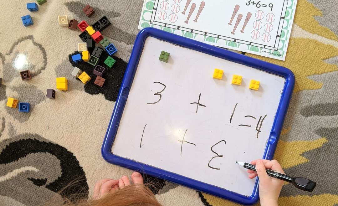 little girl writing on a white board. There is one green lego and 3 yellow legos, and she's writing 1+3 and 3+1